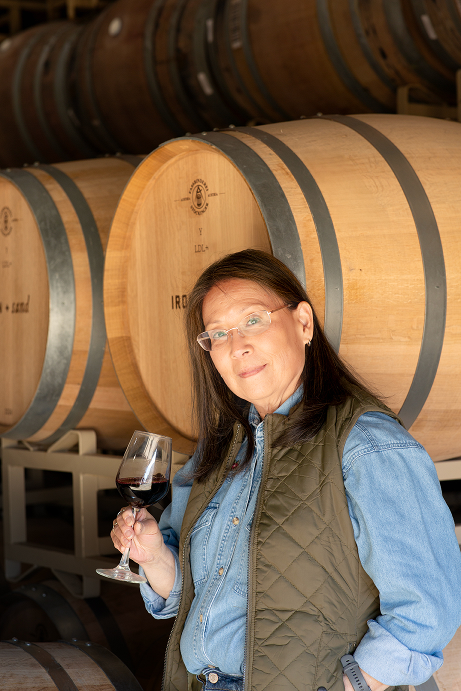 Winemaker Lee Miyamura posing with a glass of red wine in front of wine barrels